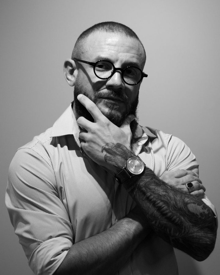 Retrato em preto e branco de Lucas Coutinho, de óculos redondos e barba, olhando para a câmera com a mão no queixo.